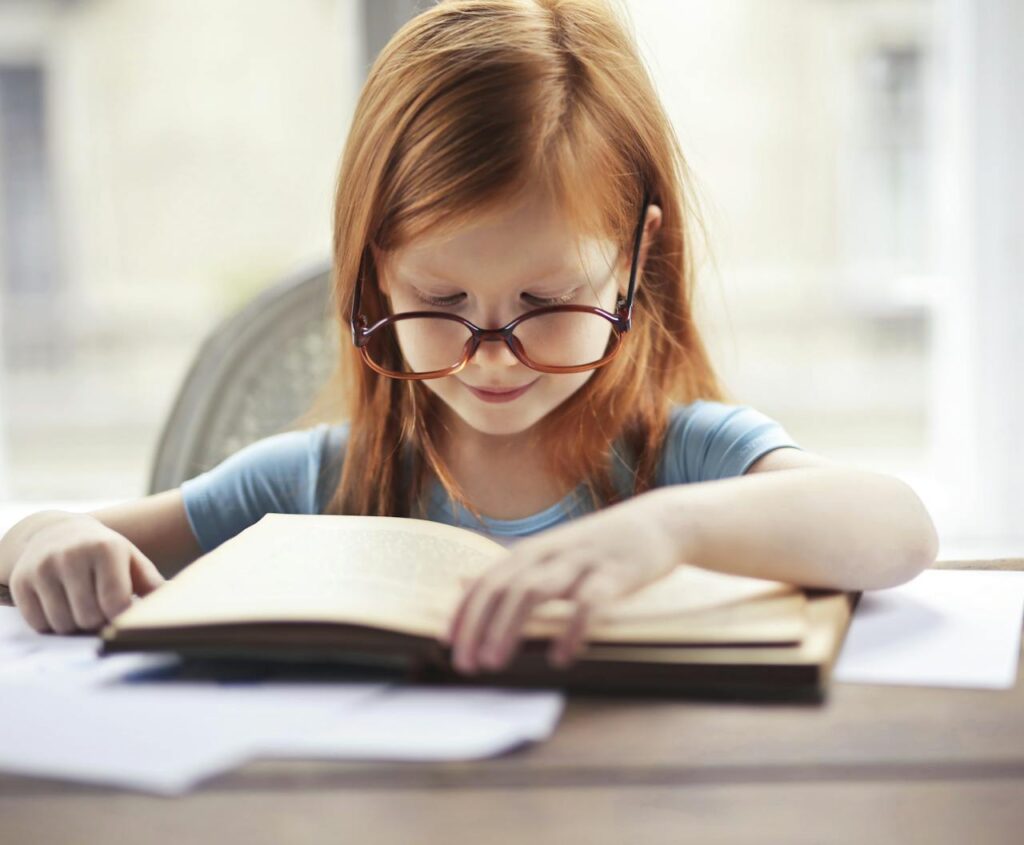 pexels-photo-3768122 A young girl with glasses intently reads a book indoors, embodying the essence of learning and discovery.